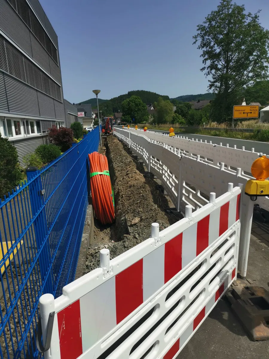 Leitungsgraben an einer Straße mit orangefarbenen Leerrohren, Absperrung und Bagger